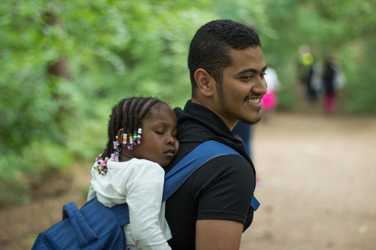 A smiling young man carrying a sleeping child on his back.