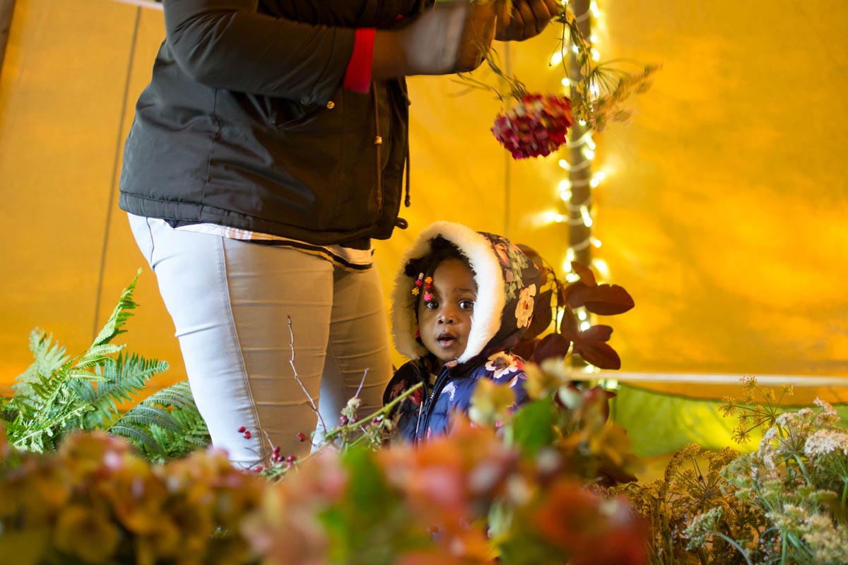 A child looks into the camera while her mother creates a garland.